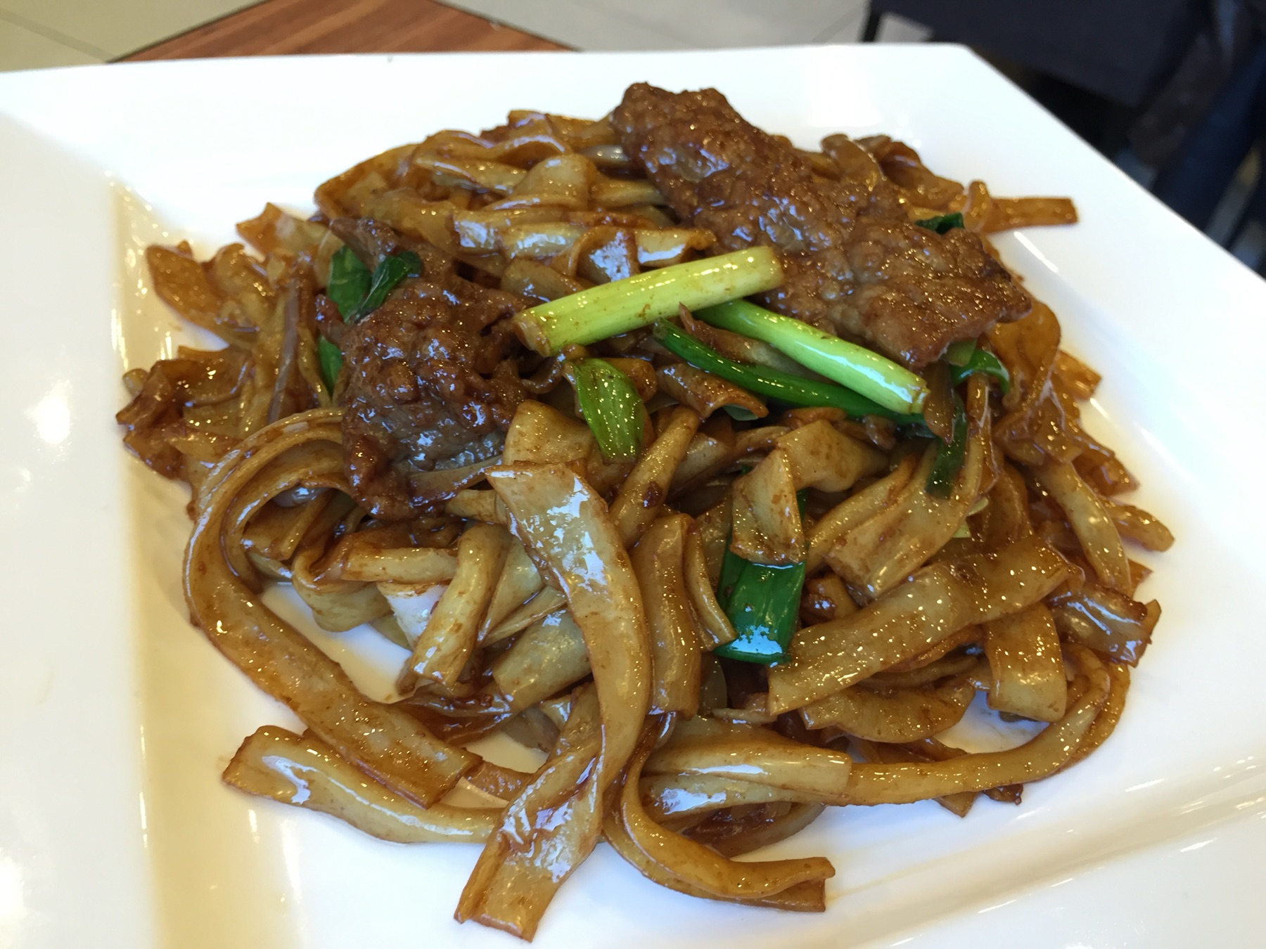 A plate of beef chow fun with glossy wide rice noodles, tender beef slices and fresh scallion batons